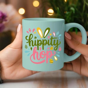 A person's hand holding a teal-colored mug with the text "hippity hop" printed on it, with a background of colorful lights and flowers.