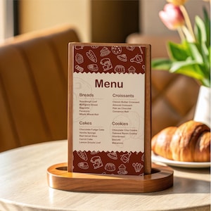 May include: A wooden menu holder displaying a menu with categories for breads, croissants, cakes, and cookies. The menu features a brown and white design with illustrations of baked goods. A croissant sits on a plate in the background.