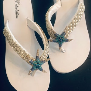 K&ouml;nnte beinhalten: Wei&szlig;e Flip-Flops mit einem Perlenriemen und einem blau-t&uuml;rkisfarbenen Seestern-Dekor. Das Wort "Redvolution" ist auf dem Fu&szlig;bett aufgedruckt. Die Flip-Flops liegen auf einer schwarzen Oberfl&auml;che.