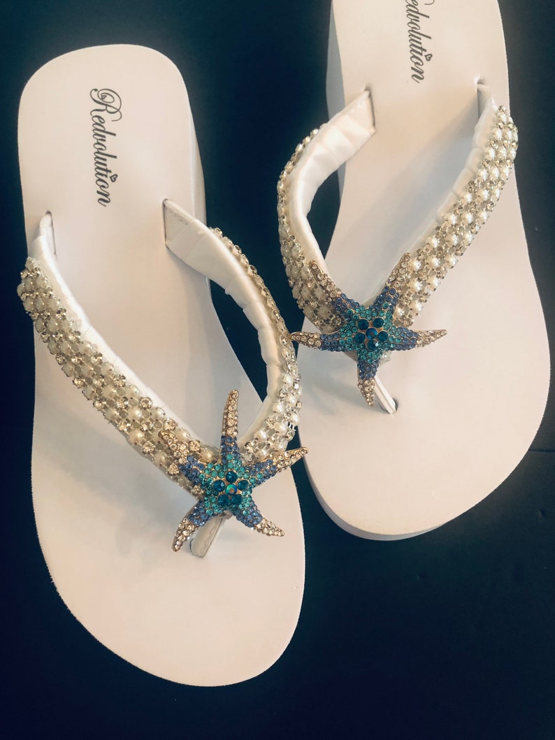 K&ouml;nnte beinhalten: Wei&szlig;e Flip-Flops mit einem Perlenriemen und einem blau-t&uuml;rkisfarbenen Seestern-Dekor. Das Wort "Redvolution" ist auf dem Fu&szlig;bett aufgedruckt. Die Flip-Flops liegen auf einer schwarzen Oberfl&auml;che.