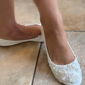 Puede incluir: Un par de bailarinas blancas con punta redondeada y detalles de encaje floral blanco. El adorno de encaje en la parte delantera de cada zapato está acentuado con pequeñas cuentas blancas redondas y lentejuelas para dar textura.