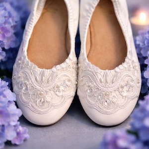 Puede incluir: Un par de bailarinas blancas con intrincados encajes superpuestos en las puntas redondeadas. El encaje está adornado con pequeñas perlas blancas y lentejuelas transparentes, creando un patrón floral texturizado. Las plantillas interiores son de color beige.