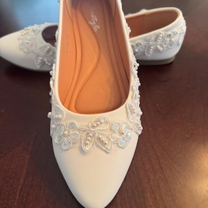 Puede incluir: Un par de zapatos planos blancos de punta afilada con superposiciones de encaje floral delicado en la punta y el talón. Los zapatos están adornados con pequeñas perlas blancas integradas en el patrón de encaje, con un forro interior color canela.