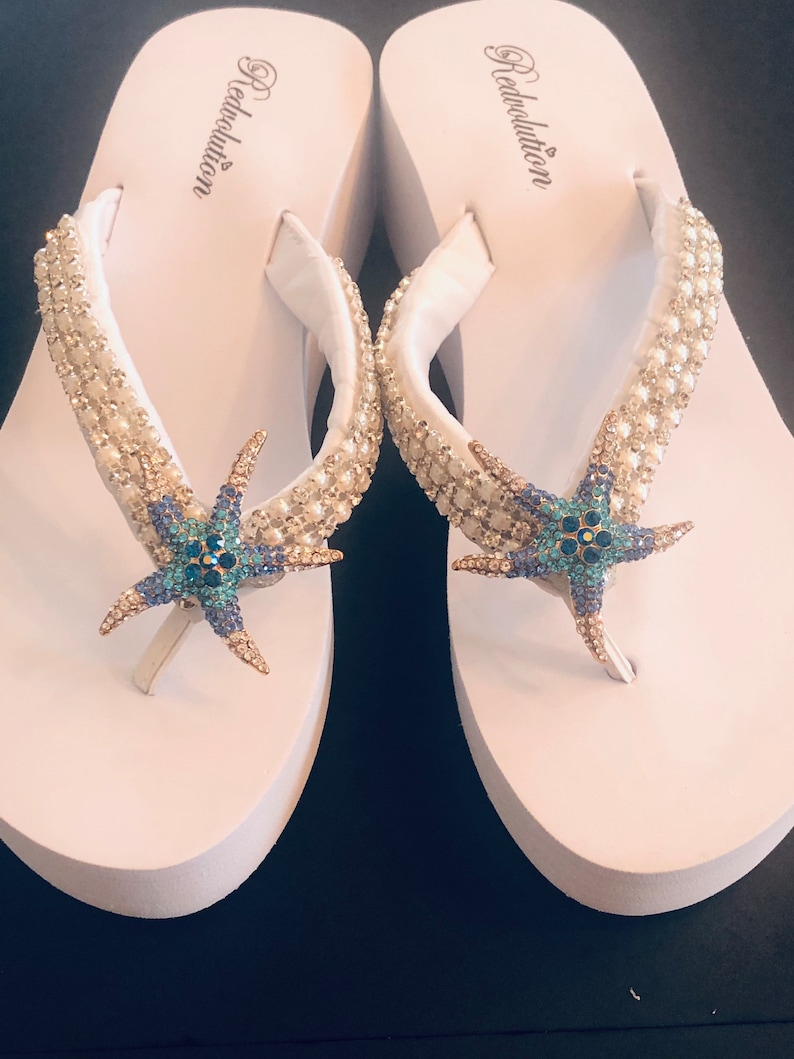 K&ouml;nnte beinhalten: Wei&szlig;e Plateau-Flip-Flops mit verzierten Riemen und Seestern-Akzenten. Die Riemen sind mit Perlen und Strasssteinen verziert. Die Seesterne sind blau, t&uuml;rkis und goldfarben. Der Markenname "Redvolution" ist auf der Seite aufgedruckt.