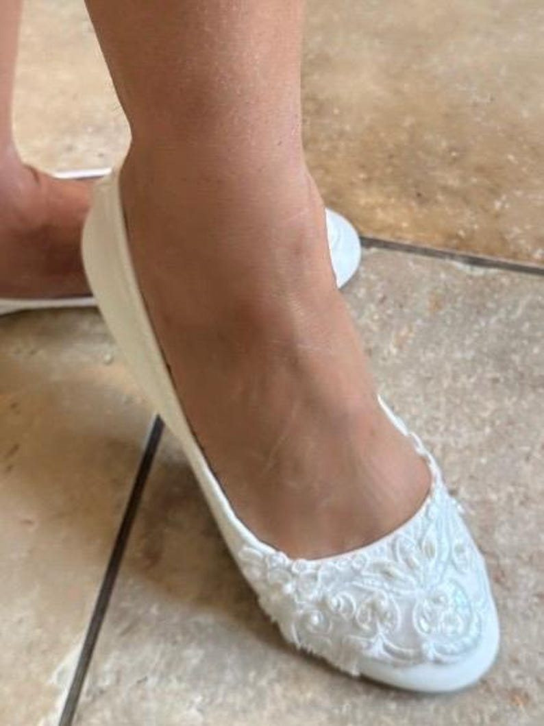 Puede incluir: Un zapato plano de color blanco con punta redondeada y un intrincado bordado de encaje floral blanco en la parte delantera. Los detalles de encaje incluyen peque&ntilde;as cuentas blancas y lentejuelas que aportan una textura y brillo sutiles.