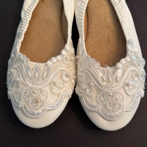 Puede incluir: Un par de bailarinas blancas con punta redondeada y suela interior color canela. El exterior est&aacute; adornado con superposiciones de encaje blanco, bordados intrincados y peque&ntilde;as perlas blancas y lentejuelas transparentes en un dise&ntilde;o floral.