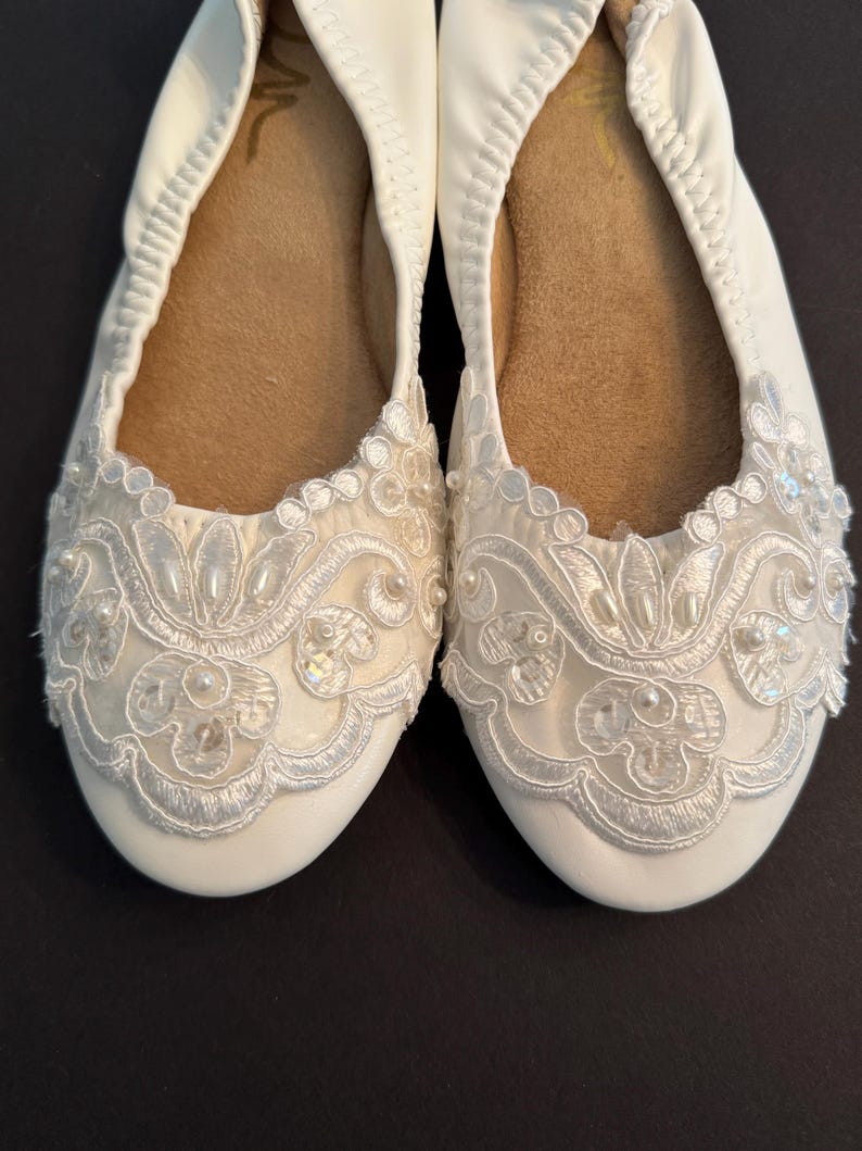 Puede incluir: Un par de bailarinas blancas con punta redondeada y suela interior color canela. El exterior est&aacute; adornado con superposiciones de encaje blanco, bordados intrincados y peque&ntilde;as perlas blancas y lentejuelas transparentes en un dise&ntilde;o floral.