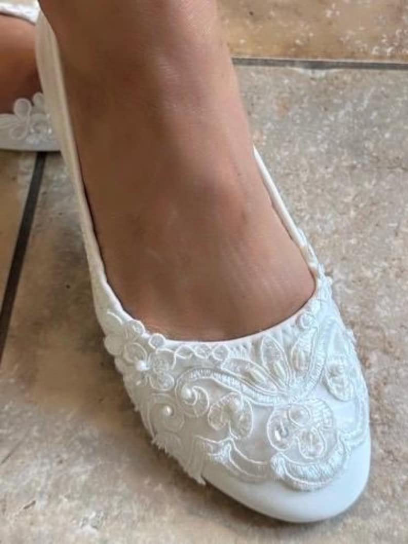 Puede incluir: Un zapato plano de color blanco que presenta un intrincado encaje superpuesto en la punta redondeada. El patr&oacute;n de encaje est&aacute; adornado con peque&ntilde;as cuentas blancas redondas y lentejuelas sutiles, creando un dise&ntilde;o texturizado al frente.