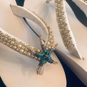K&ouml;nnte beinhalten: Wei&szlig;e Flip-Flops mit verzierten Riemen. Die Riemen sind mit klaren und perlenartigen Perlen sowie einem goldenen Seestern mit blauen und klaren Kristallen verziert. Eine stilvolle Strand-Schuh-Option.
