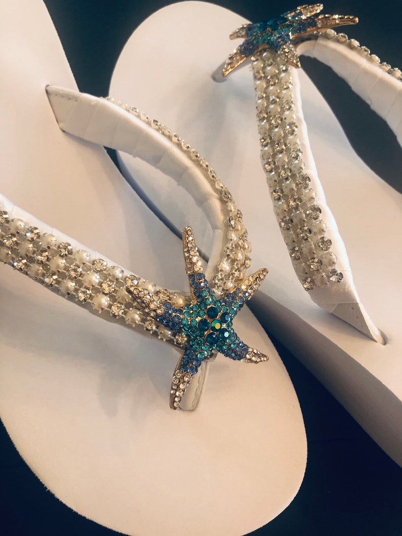 K&ouml;nnte beinhalten: Wei&szlig;e Flip-Flops mit verzierten Riemen. Die Riemen sind mit klaren und perlenartigen Perlen sowie einem goldenen Seestern mit blauen und klaren Kristallen verziert. Eine stilvolle Strand-Schuh-Option.