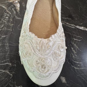 Puede incluir: Una bailarina blanca con punta redondeada y borde el&aacute;stico. La parte delantera del zapato est&aacute; adornada con un intrincado bordado de encaje blanco, lentejuelas transparentes y peque&ntilde;as perlas artificiales blancas en un dise&ntilde;o floral.