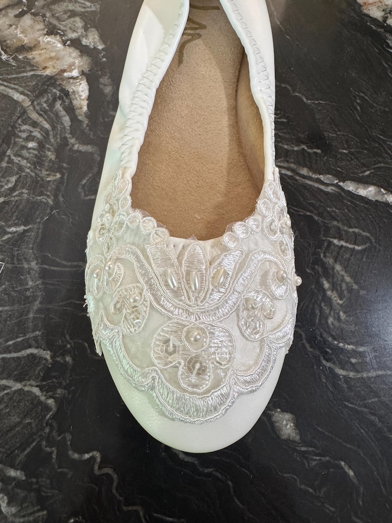 Puede incluir: Una bailarina blanca con punta redondeada y borde el&aacute;stico. La parte delantera del zapato est&aacute; adornada con un intrincado bordado de encaje blanco, lentejuelas transparentes y peque&ntilde;as perlas artificiales blancas en un dise&ntilde;o floral.
