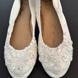 Puede incluir: Un par de bailarinas blancas con punta redondeada y borde el&aacute;stico. La parte delantera de cada zapato est&aacute; adornada con encaje floral blanco, peque&ntilde;as perlas blancas y lentejuelas transparentes. La suela interior es de color beige con talla 7.