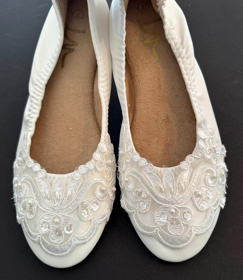 Puede incluir: Un par de bailarinas blancas con punta redondeada y borde el&aacute;stico. La parte delantera de cada zapato est&aacute; adornada con encaje floral blanco, peque&ntilde;as perlas blancas y lentejuelas transparentes. La suela interior es de color beige con talla 7.