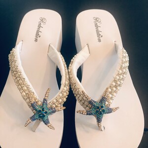 K&ouml;nnte beinhalten: Ein Paar wei&szlig;e Plateau-Flip-Flops mit Seestern-Design. Die Riemen sind mit kleinen, glitzernden Verzierungen versehen. Der Seestern ist blau und t&uuml;rkis mit goldenen Akzenten. Der Markenname "Redvolution" ist auf dem Fu&szlig;bett aufgedruckt.