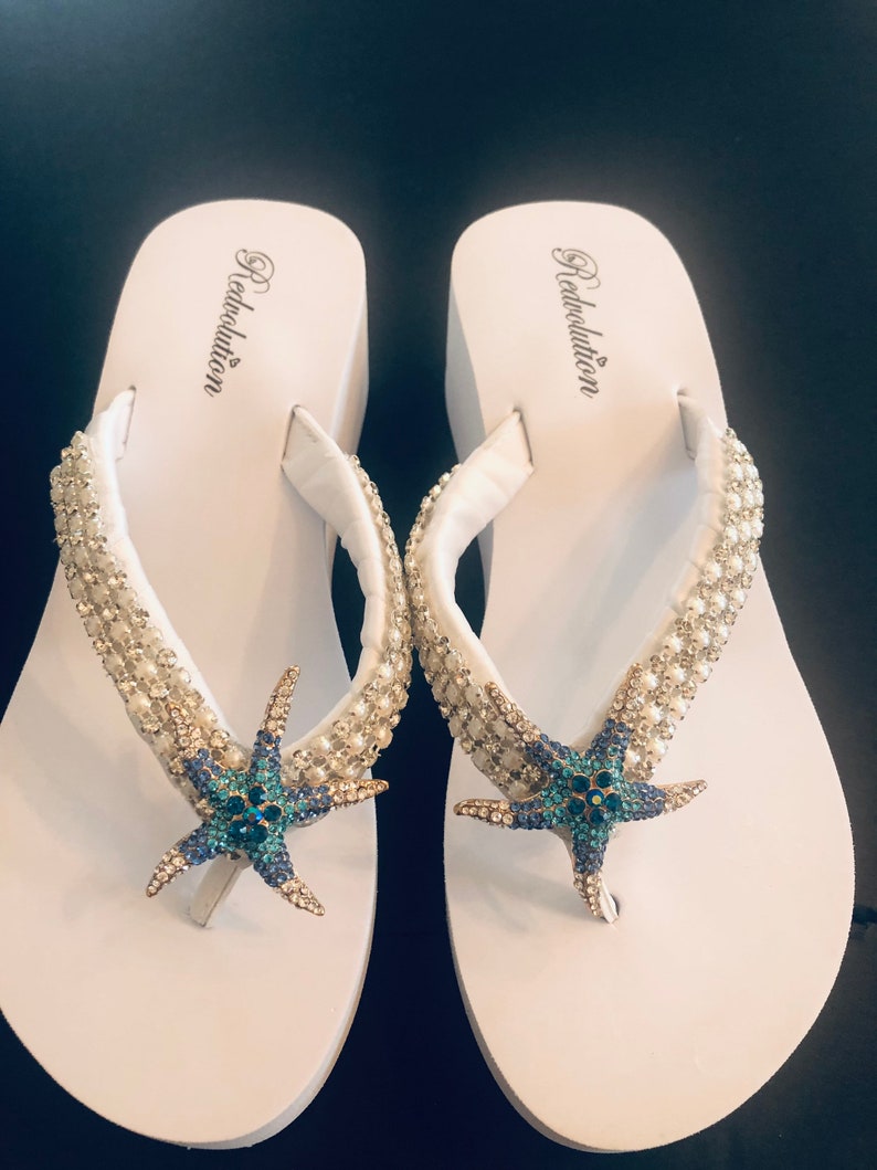 K&ouml;nnte beinhalten: Ein Paar wei&szlig;e Plateau-Flip-Flops mit Seestern-Design. Die Riemen sind mit kleinen, glitzernden Verzierungen versehen. Der Seestern ist blau und t&uuml;rkis mit goldenen Akzenten. Der Markenname "Redvolution" ist auf dem Fu&szlig;bett aufgedruckt.