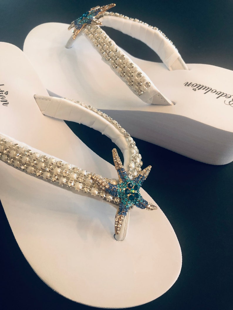 K&ouml;nnte beinhalten: Wei&szlig;e Flip-Flops mit verzierten Riemen. Die Riemen sind mit klaren und perlenartigen Perlen sowie einem blauen und goldenen Seestern verziert. Die Flip-Flops haben eine wei&szlig;e Sohle und einen Zehenriemen.