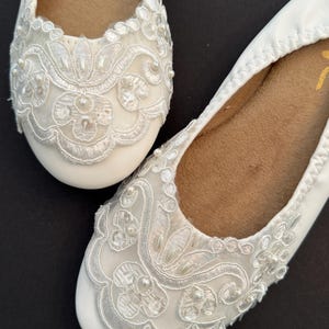 Puede incluir: Un par de bailarinas blancas con puntera redondeada y forro interior color canela. El exterior de cada zapato est&aacute; decorado con bordados de encaje floral blanco, costuras intrincadas y peque&ntilde;as cuentas blancas en la puntera.