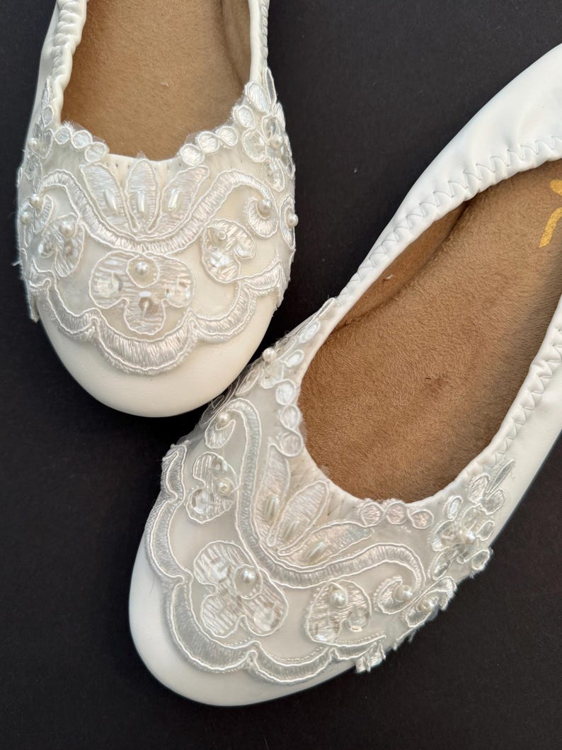 Puede incluir: Un par de bailarinas blancas con puntera redondeada y forro interior color canela. El exterior de cada zapato est&aacute; decorado con bordados de encaje floral blanco, costuras intrincadas y peque&ntilde;as cuentas blancas en la puntera.