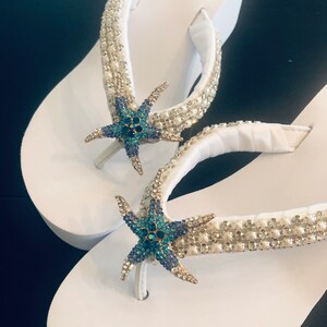 K&ouml;nnte beinhalten: Wei&szlig;e Plateau-Flip-Flops mit Strass- und Perlenbesatz. Jede Flip-Flop hat einen blau-t&uuml;rkisfarbenen, geschm&uuml;ckten Seestern. Der Markenname "Revolution" ist auf der Seite der Flip-Flops aufgedruckt.