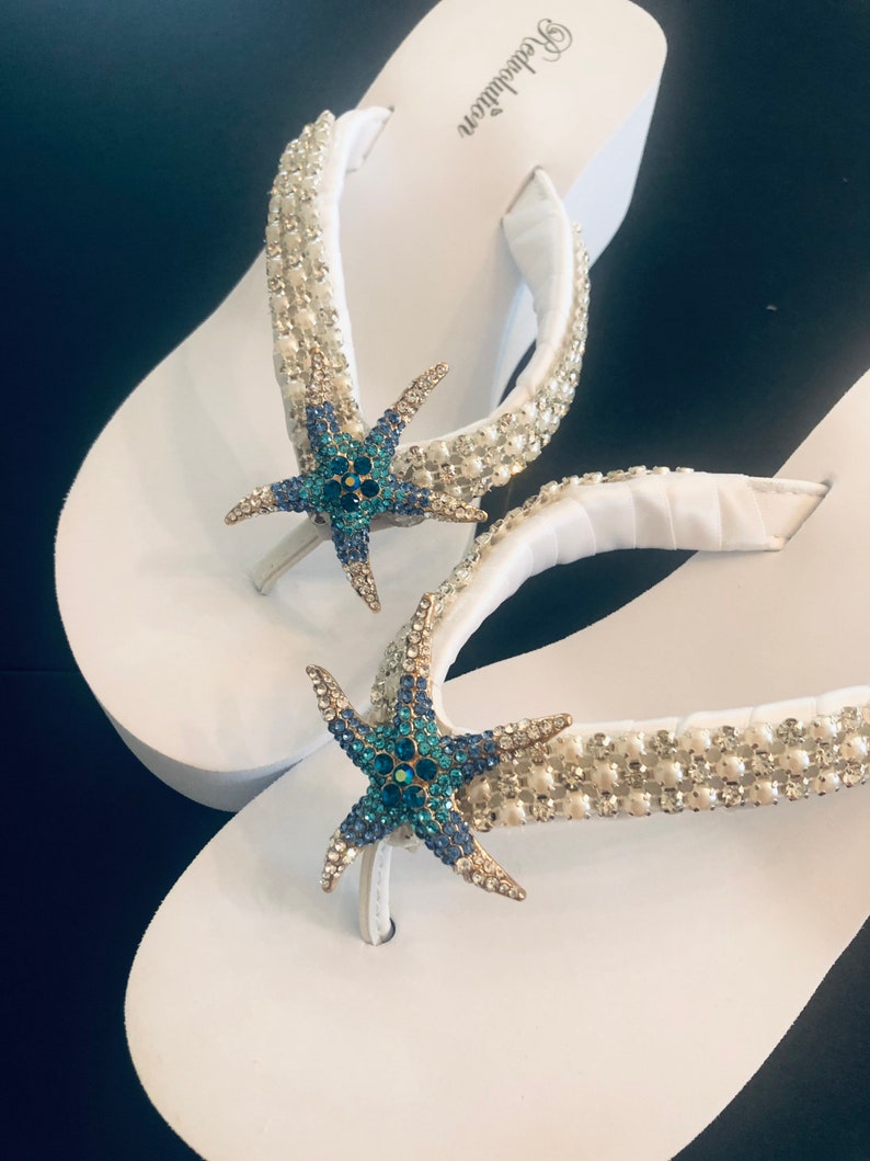 K&ouml;nnte beinhalten: Wei&szlig;e Plateau-Flip-Flops mit Strass- und Perlenbesatz. Jede Flip-Flop hat einen blau-t&uuml;rkisfarbenen, geschm&uuml;ckten Seestern. Der Markenname "Revolution" ist auf der Seite der Flip-Flops aufgedruckt.