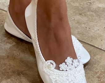 May include: A pair of white ballet flats featuring a rounded toe and a smooth finish. The front of each shoe is adorned with a white lace appliqué in a floral pattern, accented with small white pearls and intricate embroidery on a sheer mesh base.