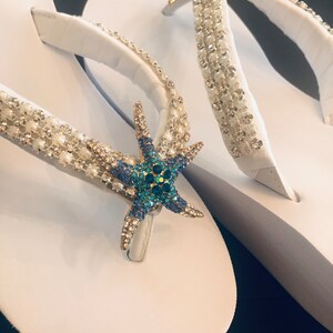 K&ouml;nnte beinhalten: Wei&szlig;e Flip-Flops mit verzierten Riemen und einem Seestern-Design. Die Riemen sind mit klaren und perlenartigen Perlen verziert, und ein goldfarbener Seestern mit blauen und gr&uuml;nen Kristallen befindet sich in der Mitte.