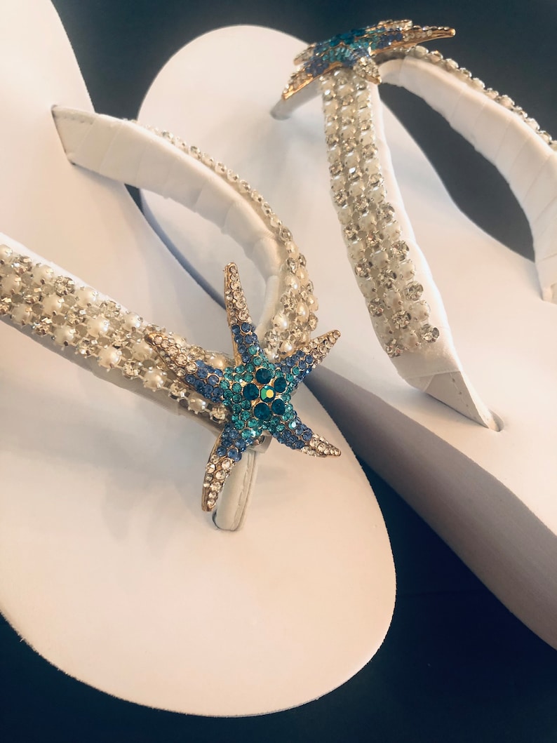 K&ouml;nnte beinhalten: Wei&szlig;e Flip-Flops mit verzierten Riemen und einem Seestern-Design. Die Riemen sind mit klaren und perlenartigen Perlen verziert, und ein goldfarbener Seestern mit blauen und gr&uuml;nen Kristallen befindet sich in der Mitte.