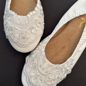 Puede incluir: Un par de bailarinas blancas con superposiciones de encaje floral blanco en las puntas redondeadas, decoradas con peque&ntilde;as cuentas y lentejuelas blancas. Las suelas interiores son de color tostado y tienen un borde el&aacute;stico fruncido.