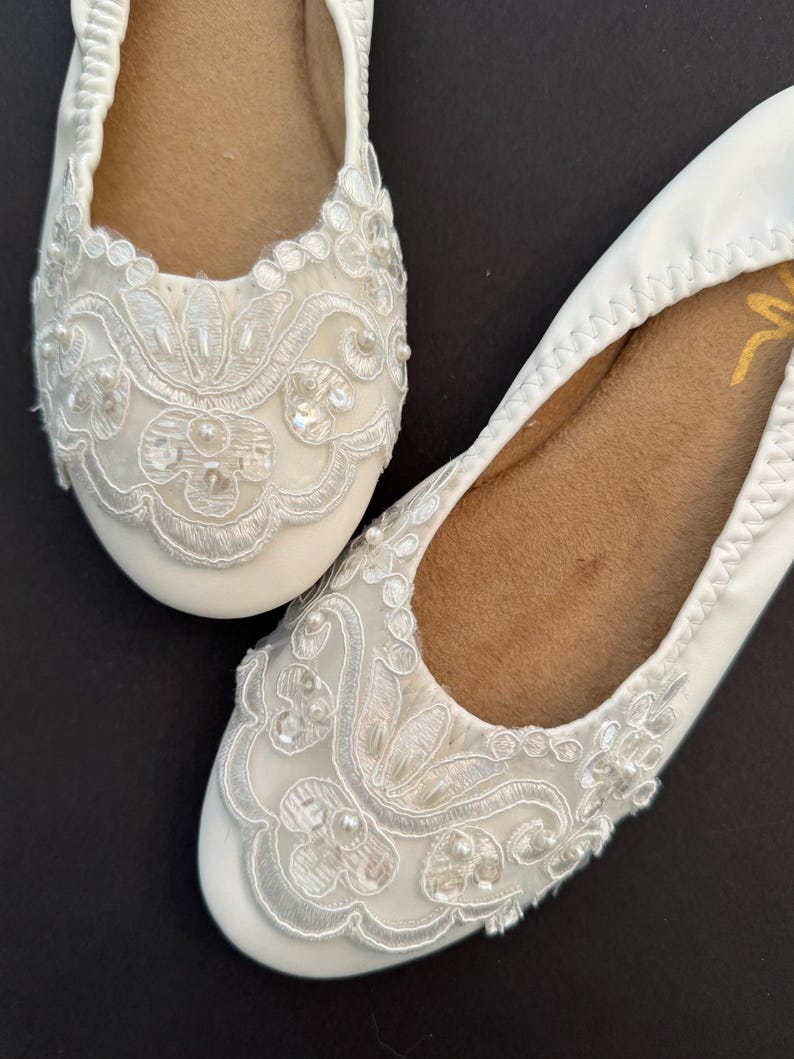 Puede incluir: Un par de bailarinas blancas con superposiciones de encaje floral blanco en las puntas redondeadas, decoradas con peque&ntilde;as cuentas y lentejuelas blancas. Las suelas interiores son de color tostado y tienen un borde el&aacute;stico fruncido.