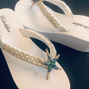 K&ouml;nnte beinhalten: Wei&szlig;e Flip-Flops mit verzierten Riemen und einem Seestern-Design. Die Riemen sind mit kleinen, funkelnden Kristallen und Perlen verziert. Ein blauer und goldener Seestern ist auf dem Riemen zentriert. Das Wort "Redolution" ist auf dem Fu&szlig;bett aufgedruckt.
