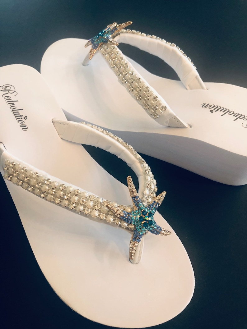 K&ouml;nnte beinhalten: Wei&szlig;e Flip-Flops mit verzierten Riemen und einem Seestern-Design. Die Riemen sind mit kleinen, funkelnden Kristallen und Perlen verziert. Ein blauer und goldener Seestern ist auf dem Riemen zentriert. Das Wort "Redolution" ist auf dem Fu&szlig;bett aufgedruckt.