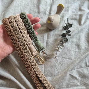Könnte beinhalten: Ein Set aus vier geflochtenen Schlüsselbändern oder Handschlaufen in verschiedenen Braun-, Beige- und Salbeigrüntönen. Jedes Band ist mit einem Metallkarabiner in Gold-, Silber- oder Schwarztönen versehen und weist ein strukturiertes Strickmuster auf.