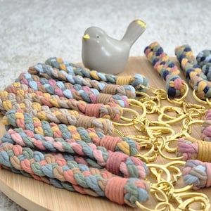 Può includere: Una collezione di portachiavi intrecciati multicolore, rosa, blu, giallo e verde, con anelli dorati. Un piccolo uccello in ceramica grigia con becco e punte delle ali dorate è posizionato vicino.