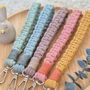Op de afbeelding: Vijf macramé polsband-sleutelhangers in pasteltinten van mintgroen, lichtblauw, lavendel, oudroze en mosterdgeel. Elke sleutelhanger heeft een gevlochten koordontwerp met een zilverkleurige metalen karabijnhaak bevestigd aan een gewikkelde basis.