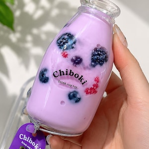 May include: A clear plastic bottle filled with a light purple liquid containing dark blue berries and red berry accents. The bottle features the black text Chiboki Drink cup toy and includes a purple tag with the same text and berry imagery.