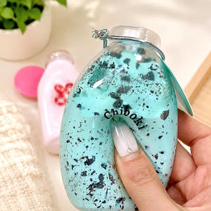 Peut inclure: Un jouet souple en forme de bouteille vert menthe, rempli de particules sombres et mouchetées, avec le texte "Chiboki" imprimé en noir. Le matériau flexible est enfoncé par la pression, révélant sa texture malléable et sa forme arrondie.
