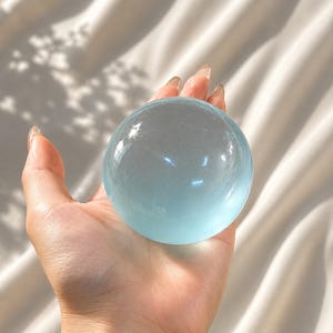 以下が含まれることがあります： 滑らかで球形の半透明なライトブルーのクリスタルボール。表面は磨かれており、光を反射して丸みを帯びた上部に明るいハイライトを作り出しています。この球体は透明で、全体的に淡い青色をしています。