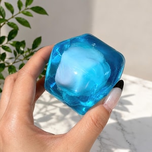 Könnte beinhalten: Ein transparentes, blaues, würfelförmiges Objekt mit einem weißen, wolkenartigen Inneren. Das Objekt wird in einer Hand gehalten, und die blaue Farbe verleiht ihm ein wässriges Aussehen. Die Kanten des Würfels sind leicht abgerundet.