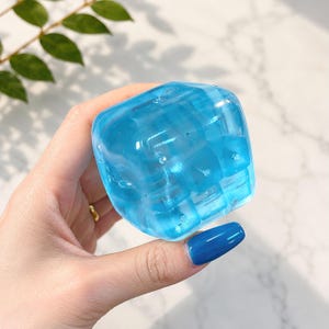 Könnte beinhalten: Ein durchscheinendes, hellblaues, würfelförmiges Objekt. Der Würfel hat ein facettiertes Design, das ihm ein mehrdimensionales Aussehen verleiht. Das Objekt wird von einer Hand mit einem goldenen Ring und blauem Nagellack gehalten.