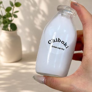 May include: A small, clear glass bottle filled with a white liquid, featuring a rounded shape and a narrow neck. The front of the bottle displays the black text "Chiboki" above the smaller text "Drink cup toy" printed in a simple font.