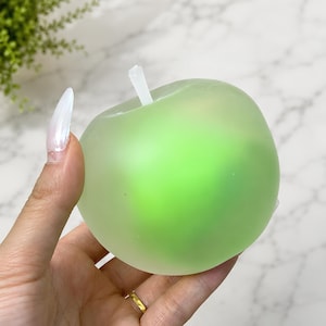 May include: A translucent, light green apple-shaped object with a smooth, matte finish and a small, white, cylindrical stem protruding from the top. The object has a rounded, bulbous form and a soft, semi-opaque appearance throughout its surface.