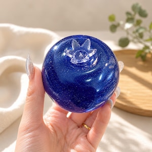 Può includere: Un oggetto sferico in resina blu intenso a forma di mirtillo, caratterizzato da una finitura liscia e lucida con particelle di glitter incorporate e un calice in plastica trasparente a forma di stella fissato sulla parte superiore.