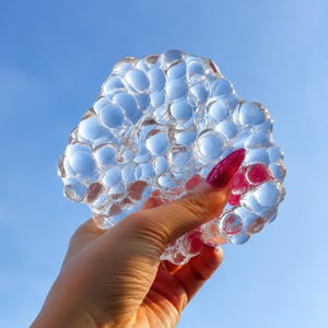 May include: A clear, transparent, and malleable substance with a textured surface composed of numerous rounded, bubble-like protrusions. The material is held up against a bright blue sky, allowing light to pass through its glossy, irregular form.