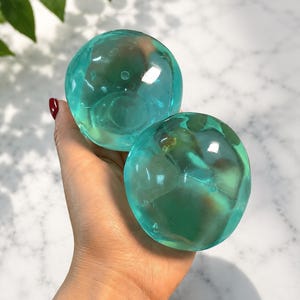 Könnte beinhalten: Zwei klare, türkisfarbene Kugeln werden in einer Hand gehalten. Die Kugeln haben eine glatte, abgerundete Form und scheinen aus Glas oder einem ähnlichen transparenten Material zu bestehen. Der Hintergrund ist eine helle Oberfläche mit ein paar grünen Blättern.