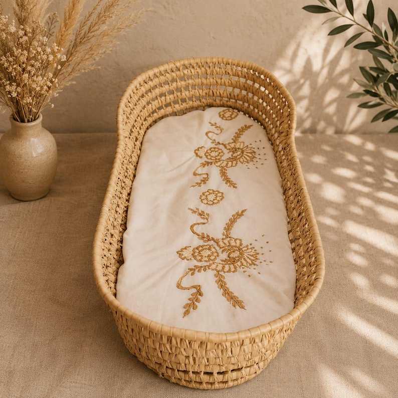 Puede incluir: Una cuna de mimbre tejido con un forro bordado de color crema. El forro presenta un dise&ntilde;o floral dorado. La cuna es de forma ovalada y tiene un color marr&oacute;n claro natural. La imagen est&aacute; tomada desde arriba.