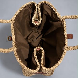 May include: A top-down view of a woven straw tote bag featuring a natural tan braided exterior and a solid dark brown fabric lining. The bag includes two braided straw handles, a small brown leather side tab, and a gold-toned metal buckle detail.