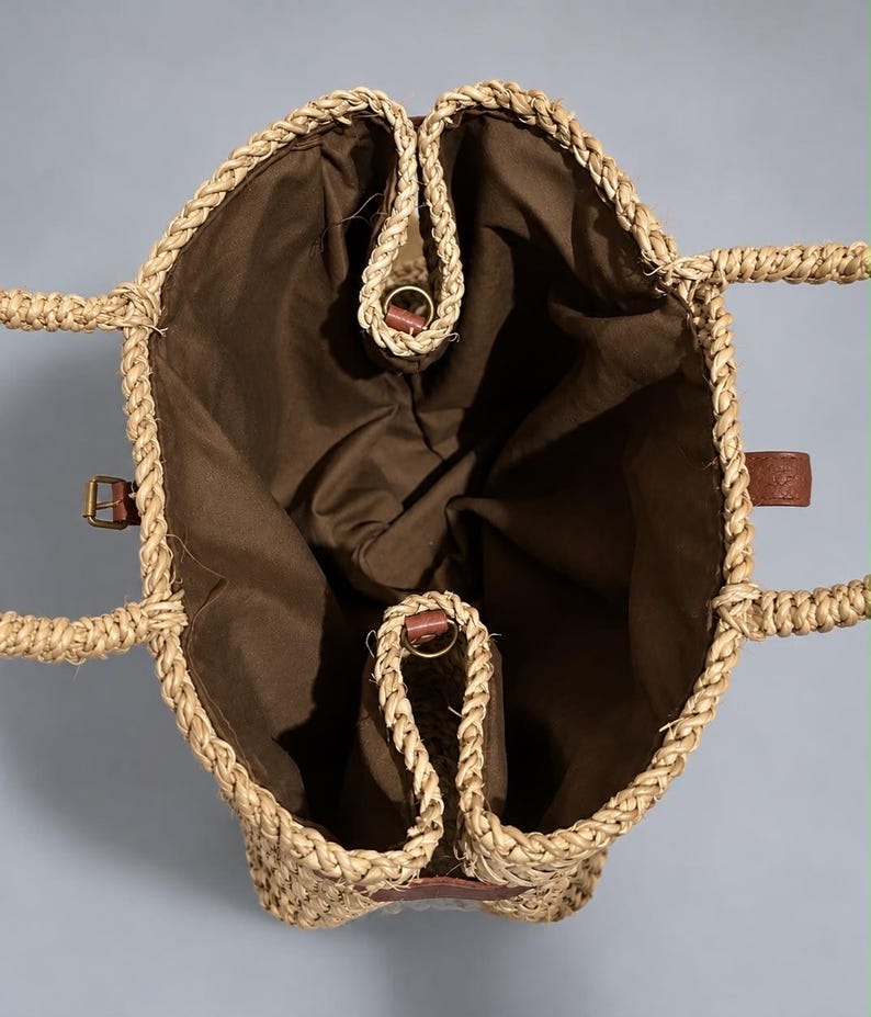 May include: A top-down view of a woven straw tote bag featuring a natural tan braided exterior and a solid dark brown fabric lining. The bag includes two braided straw handles, a small brown leather side tab, and a gold-toned metal buckle detail.