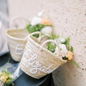 May include: Woven straw basket with white embroidered name "Elizabeth". The basket has a round handle and is filled with white and peach flowers. Another basket with the letter "E" is visible in the background.