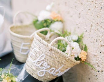 May include: Woven straw basket with white embroidered name "Elizabeth". The basket has a round handle and is filled with white and peach flowers. Another basket with the letter "E" is visible in the background.